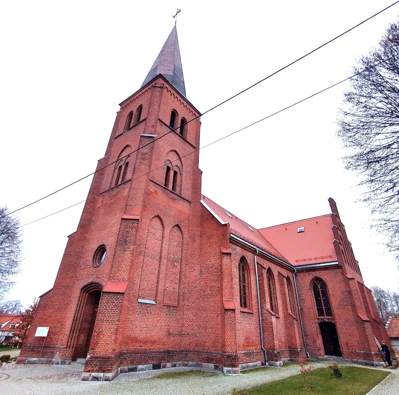 Ceglany, neogotycki kościół z wysoką wieżą z ostrą iglicą, otoczony drzewami na tle jasnego nieba