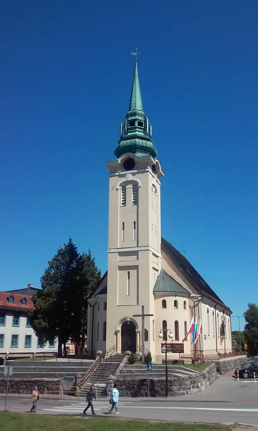 Jasna wieża kościoła z zielonym, miedzianym hełmem wznosi się nad białą nawą i schodami przy ulicy, na tle głębokiego błękitnego nieba