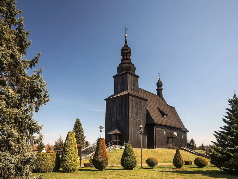 Czarny, drewniany kościół w Gwoździanach z wieżą zwieńczoną cebulastym hełmem, otoczony ogrodem