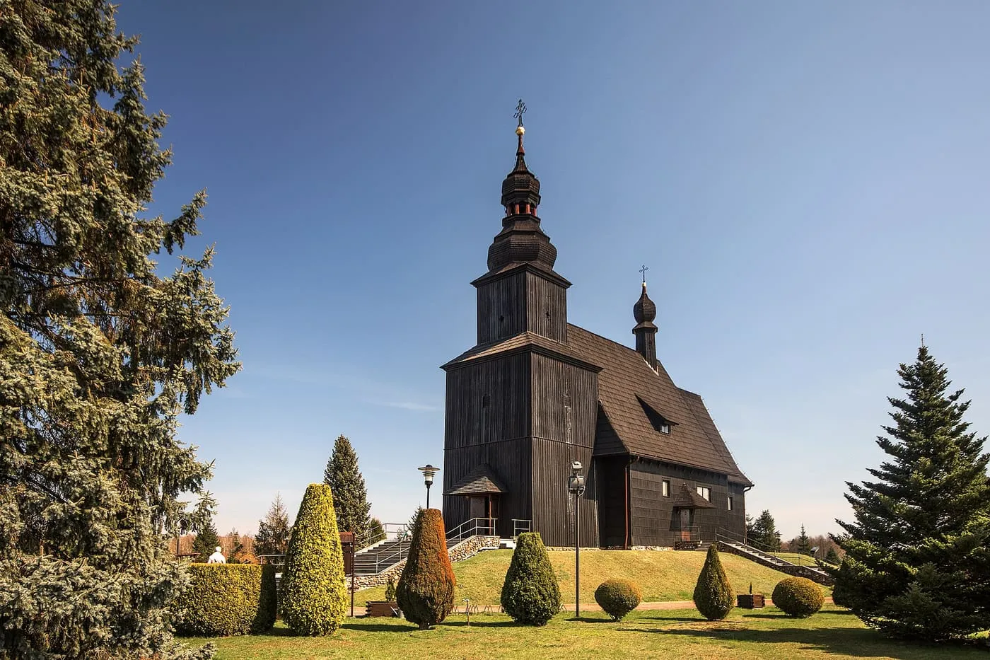 Czarny, drewniany kościół w Gwoździanach z wieżą zwieńczoną cebulastym hełmem, otoczony ogrodem