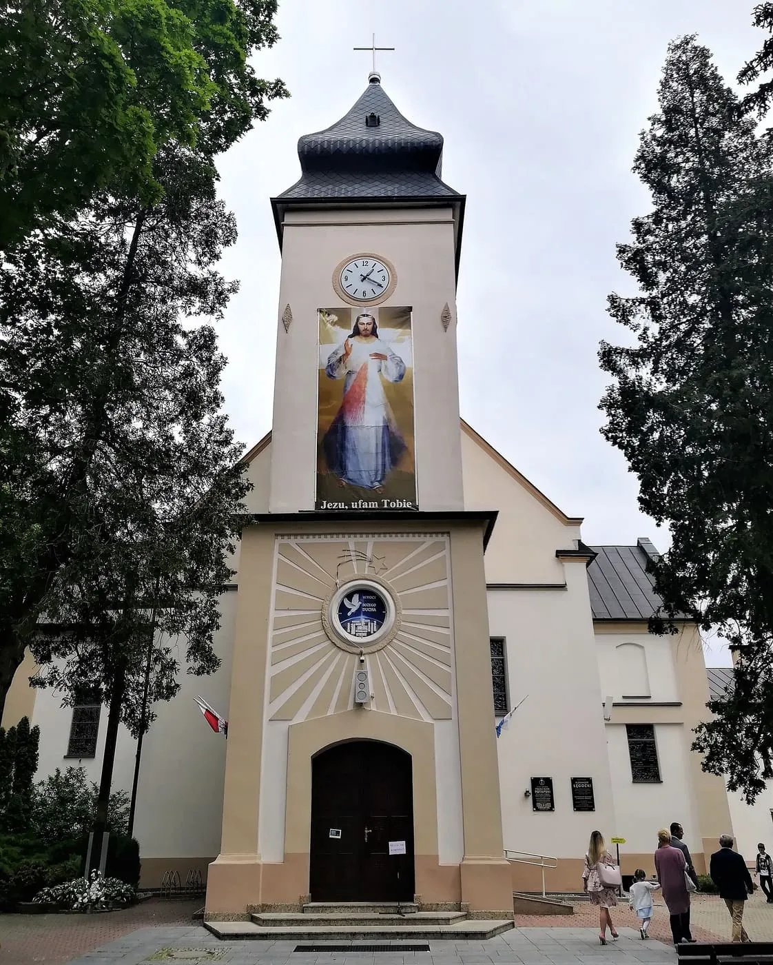 Kremowa wieża kościoła Narodzenia NMP w Bełchatowie z ciemnym, spiczastym hełmem, zegarem i banerem obrazu Jezu, ufam Tobie, przy której gromadzą się parafianie