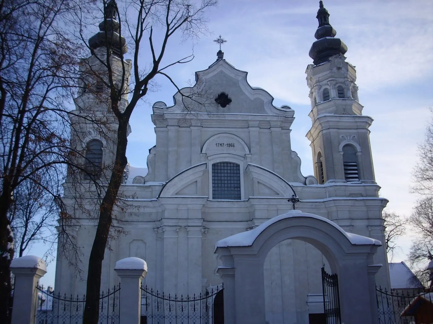 Biała, późnobarokowa fasada kościoła z dwiema wieżami zwieńczonymi ciemnymi hełmami; na pierwszym planie zaśnieżona brama z łukiem i bezlistne drzewa