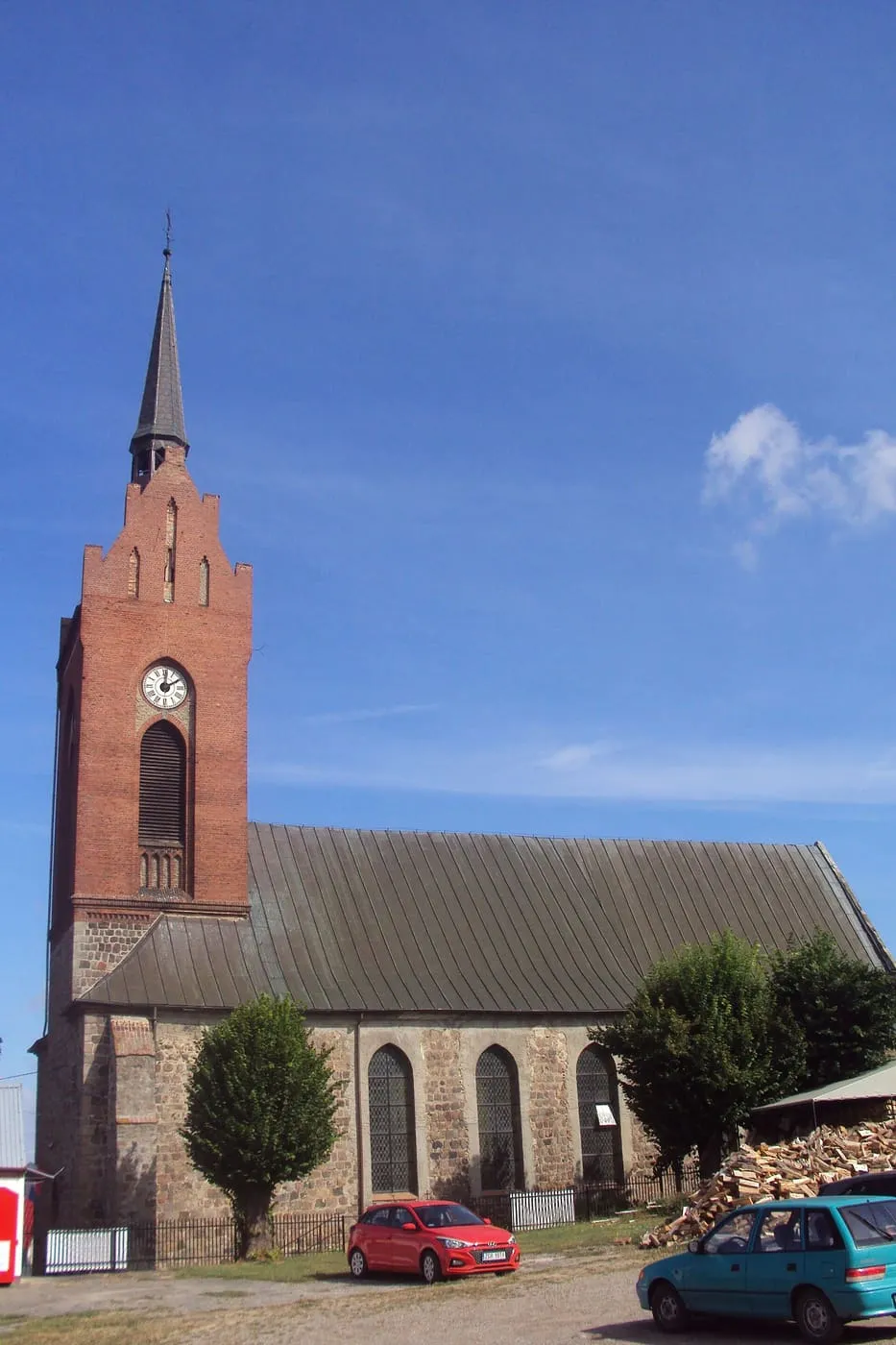 Kościół z nawą z granitowych ciosów i ceglaną wieżą z zegarem i iglicą, przed nim zaparkowane samochody