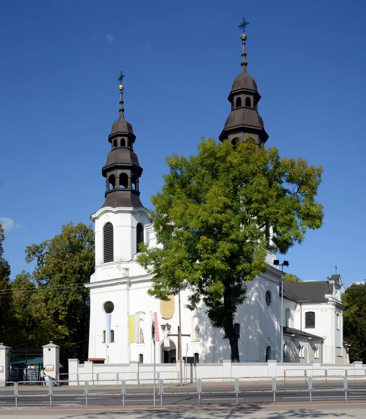 Biały, neobarokowy kościół Narodzenia NMP w Mińsku Mazowieckim z dwiema wieżami zwieńczonymi ciemnymi, wielokondygnacyjnymi hełmami i papieskimi flagami na fasadzie