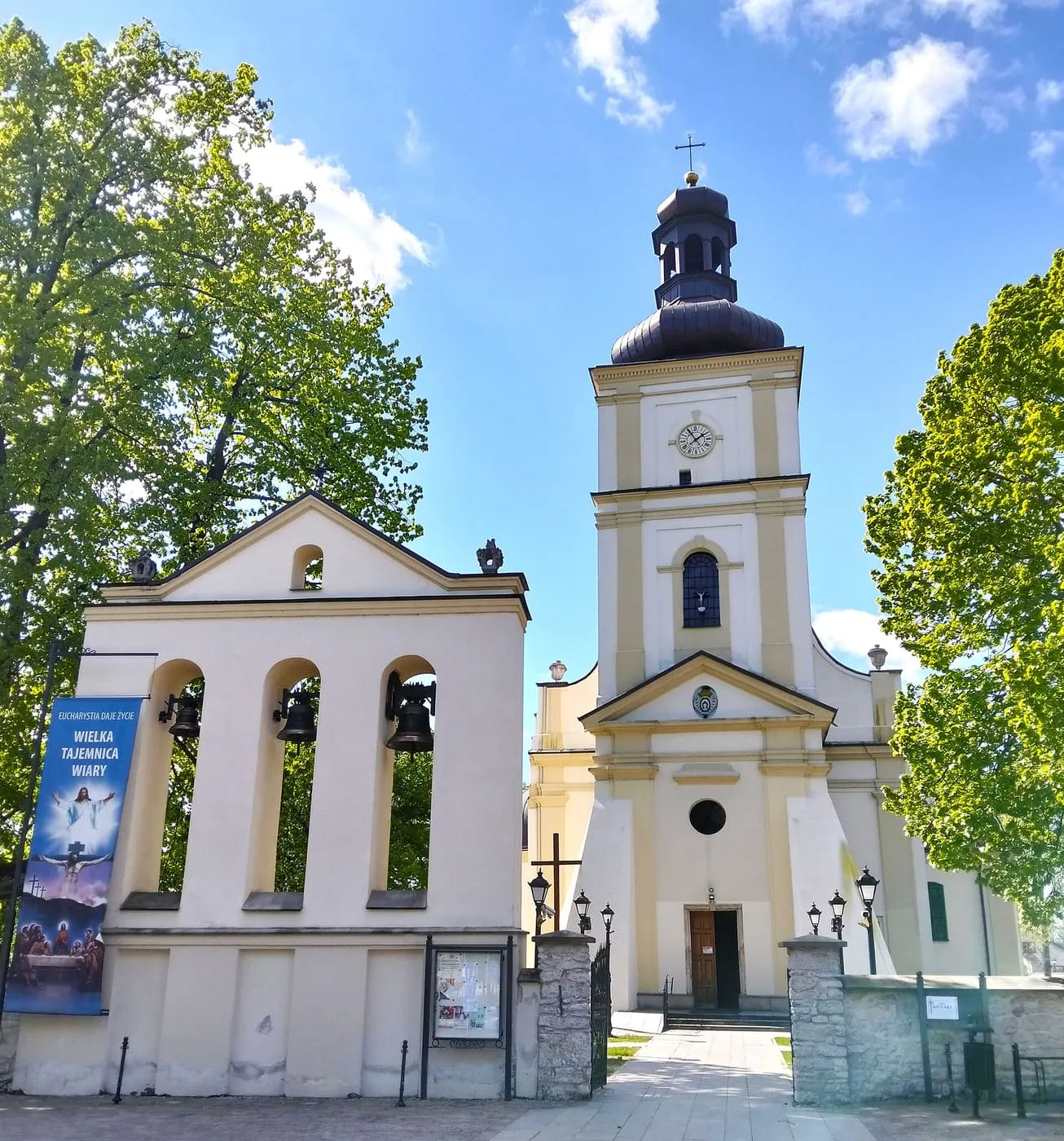 Biała dzwonnica-brama z trzema dzwonami i kościół z dwukondygnacyjną wieżą zwieńczoną kopułką, wśród zielonych drzew