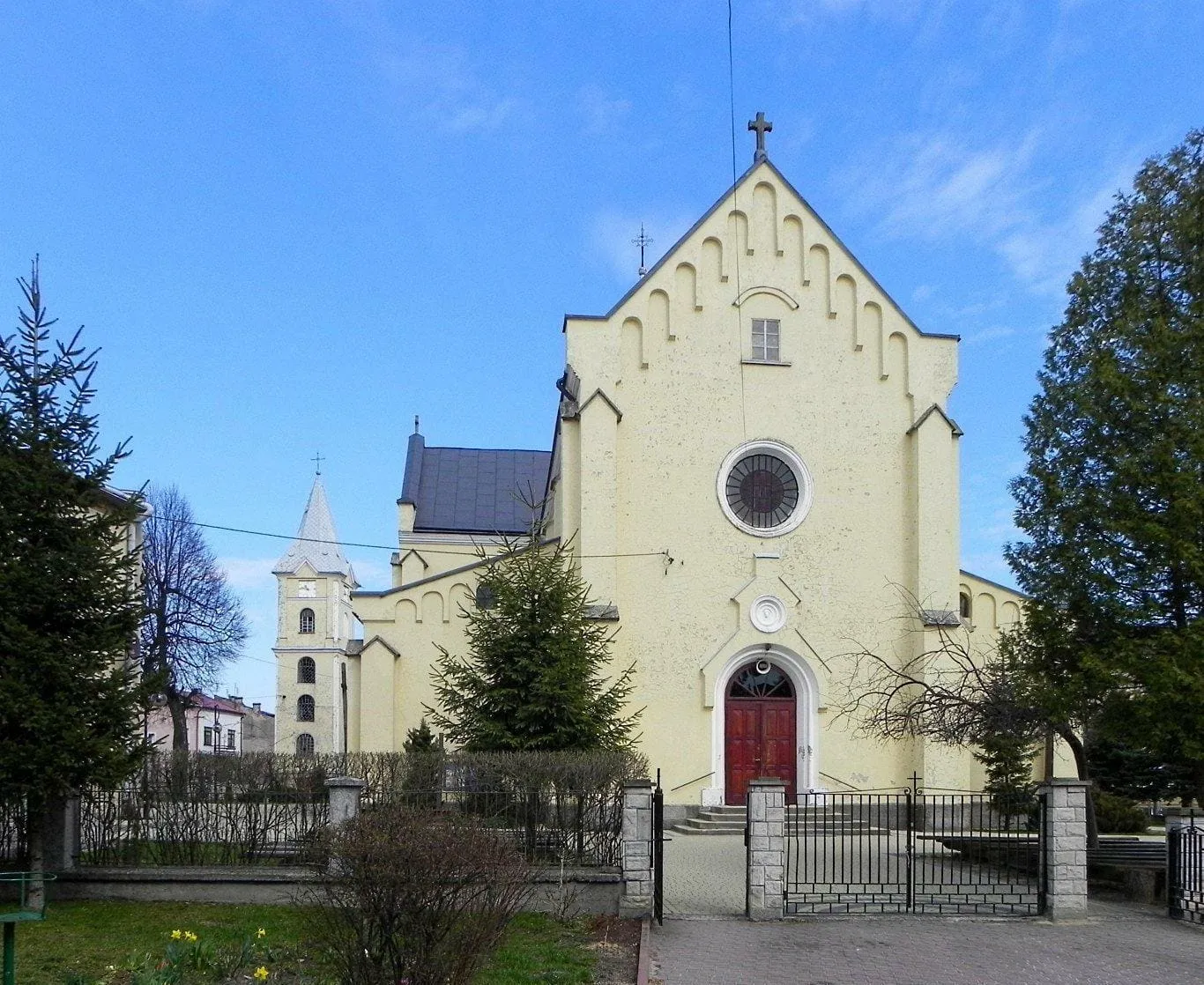 Żółta fasada neogotyckiego kościoła ze schodkowym szczytem, rozetą i czerwonymi drzwiami, za kutym ogrodzeniem, z białą wieżą w tle, pod błękitnym niebem