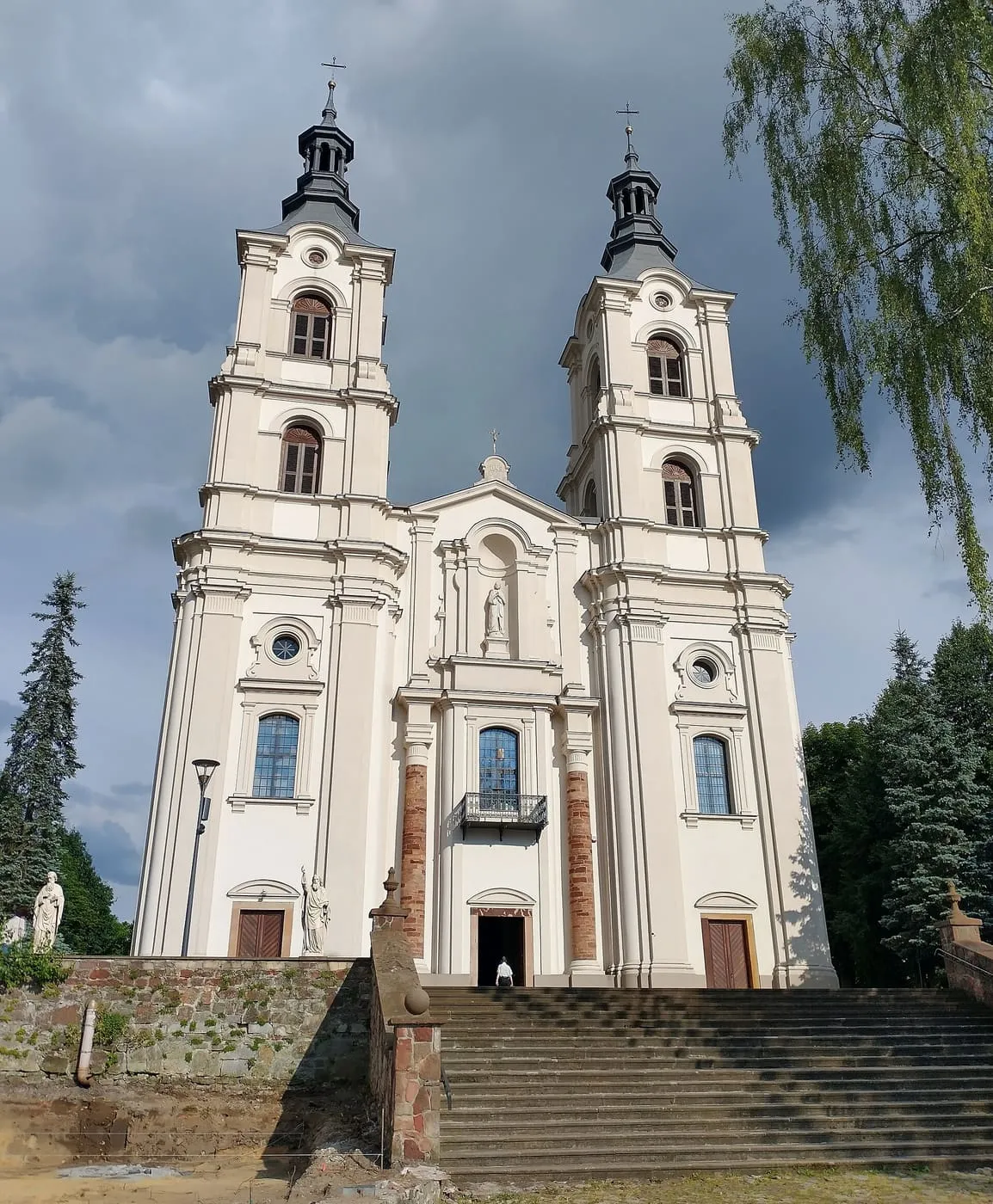 Biała, barokowa fasada kościoła z dwiema wieżami z ciemnymi hełmami, do której prowadzą szerokie schody, pod pochmurnym niebem