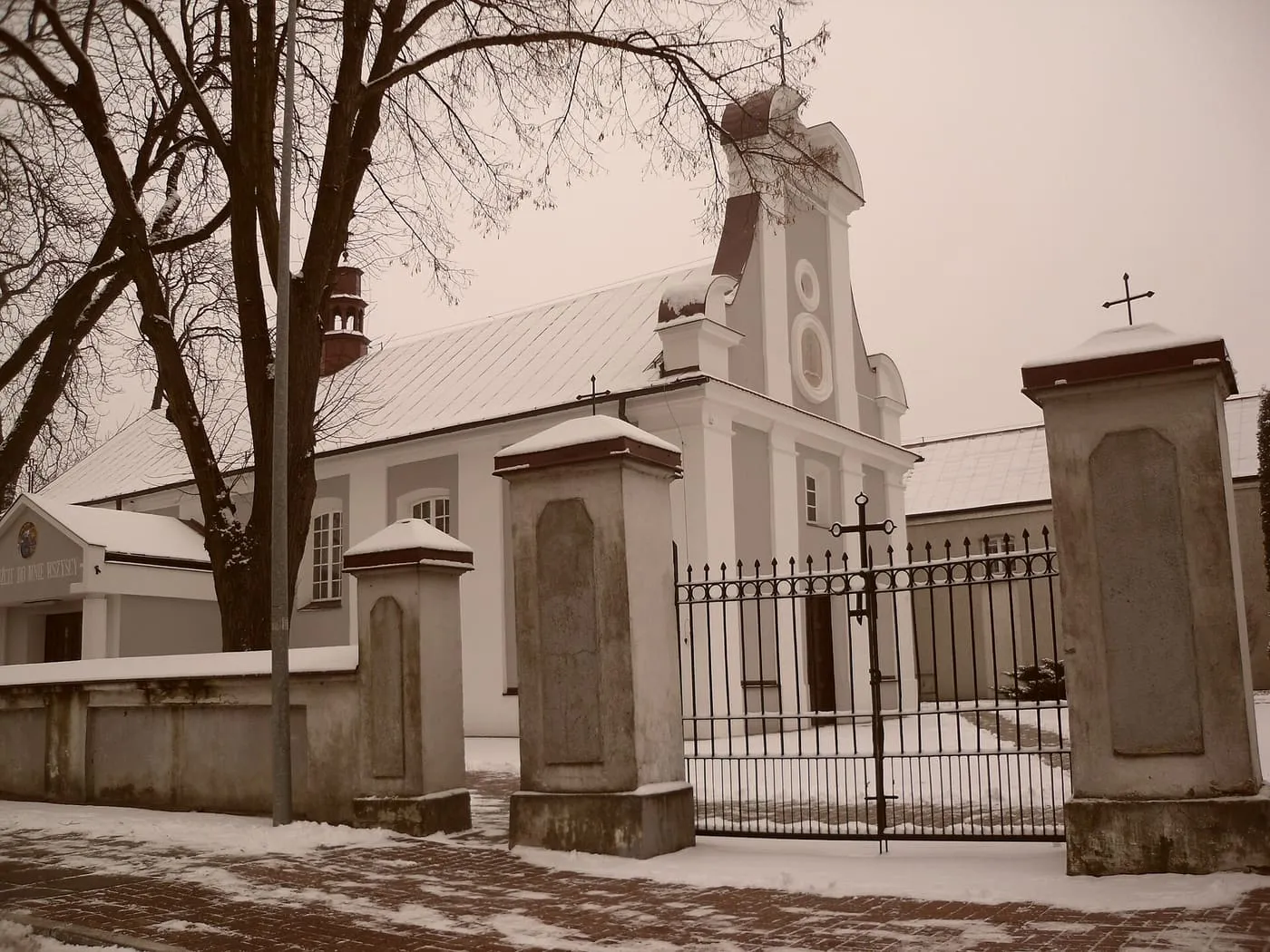 Zaśnieżony kościół o białej, późnobarokowej fasadzie ze szczytem z wolutami, widziany zza żelaznej bramy między kamiennymi słupami, wśród bezlistnych drzew