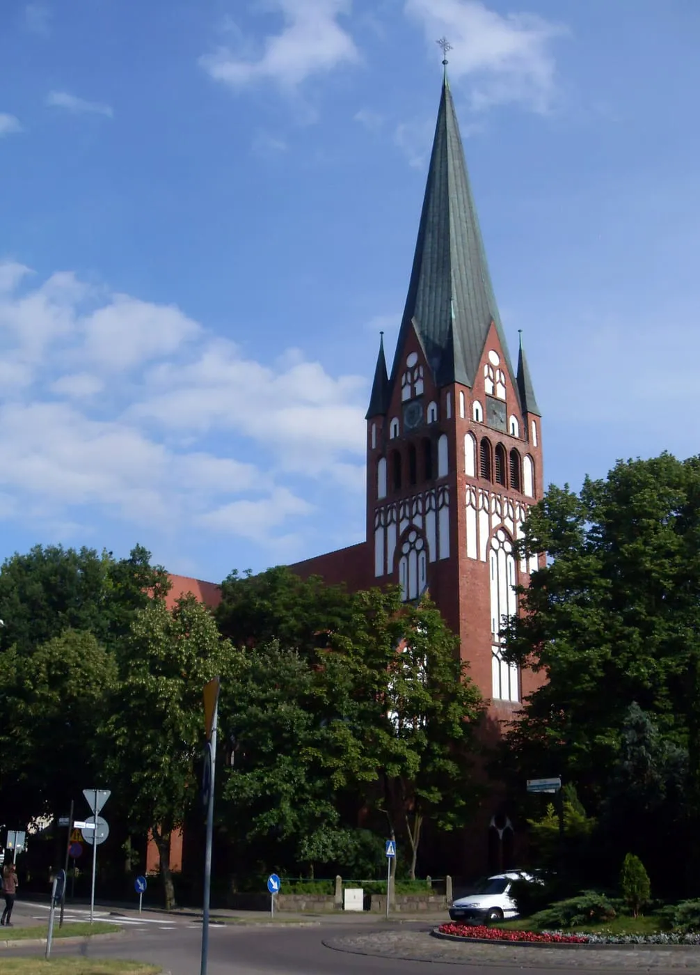 Ceglany, neogotycki kościół w Szczecinku z 78-metrową wieżą zwieńczoną smukłą, zieloną iglicą i tynkowanymi blendami, widziany zza drzew od ronda