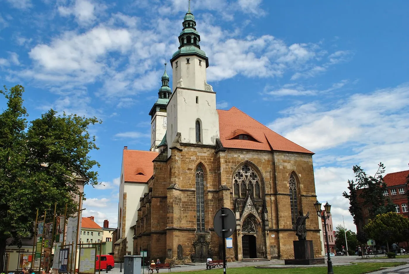 Gotycki kościół Narodzenia NMP w Złotoryi z piaskowca, z dużym oknem maswerkowym nad ostrołukowym portalem i białą wieżą zwieńczoną zielonym, wielokondygnacyjnym hełmem, od strony placu
