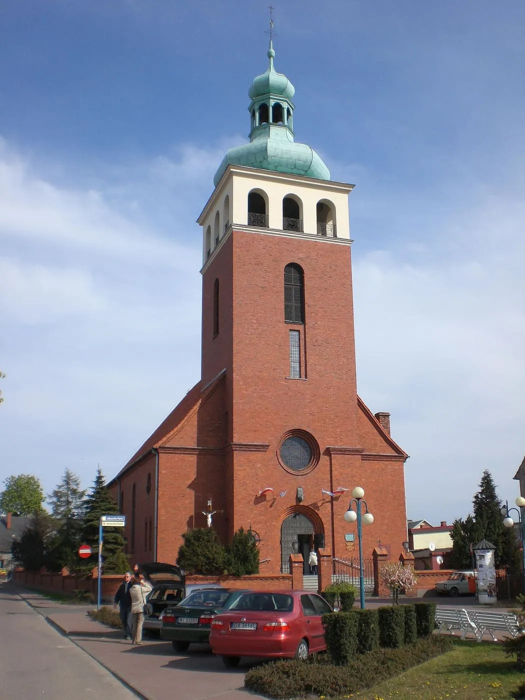 Ceglany kościół z wysoką wieżą zwieńczoną zielonkawą kopułą z latarnią, przed którym stoją zaparkowane samochody, na tle błękitnego nieba