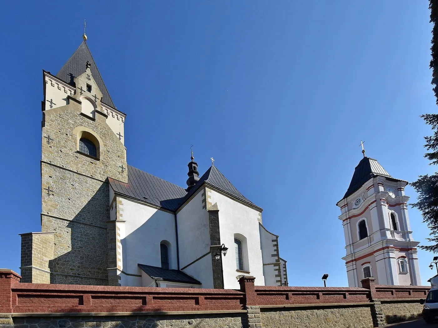 Kamienna, gotycka wieża kościoła w Lesku z neogotycką iglicą, obok osobno stojącej różowo-białej barokowej dzwonnicy z zegarem