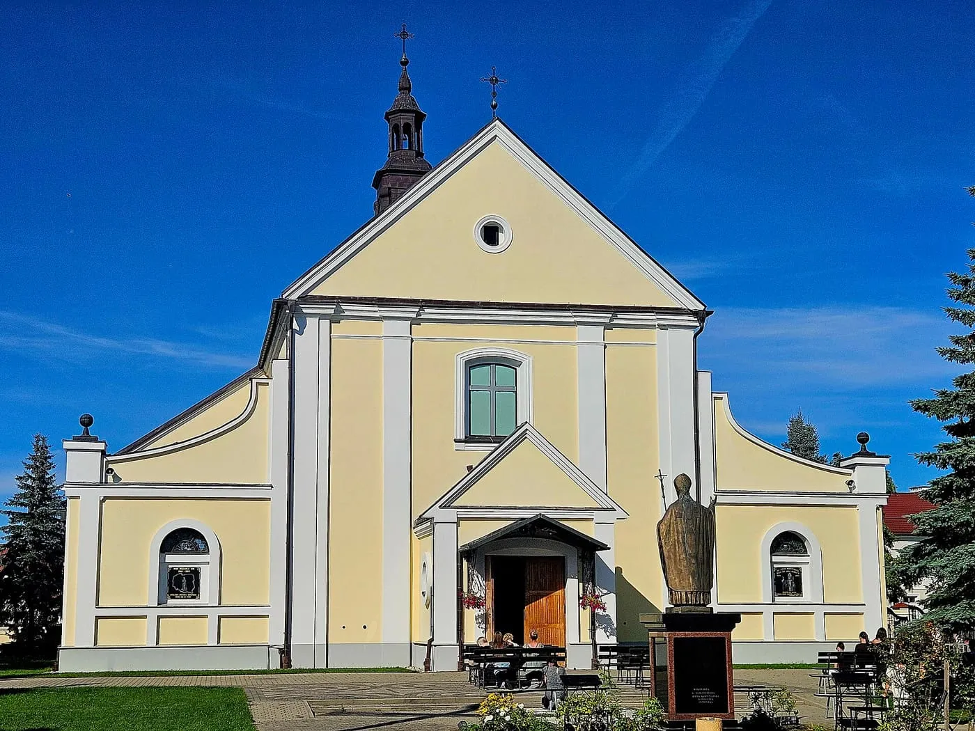 Jasnożółta fasada kościoła Nawiedzenia NMP w Ostrołęce z trójkątnym szczytem, białymi pilastrami i ciemną wieżyczką na dachu, przed nią brązowy pomnik duchownego i ławki z wiernymi