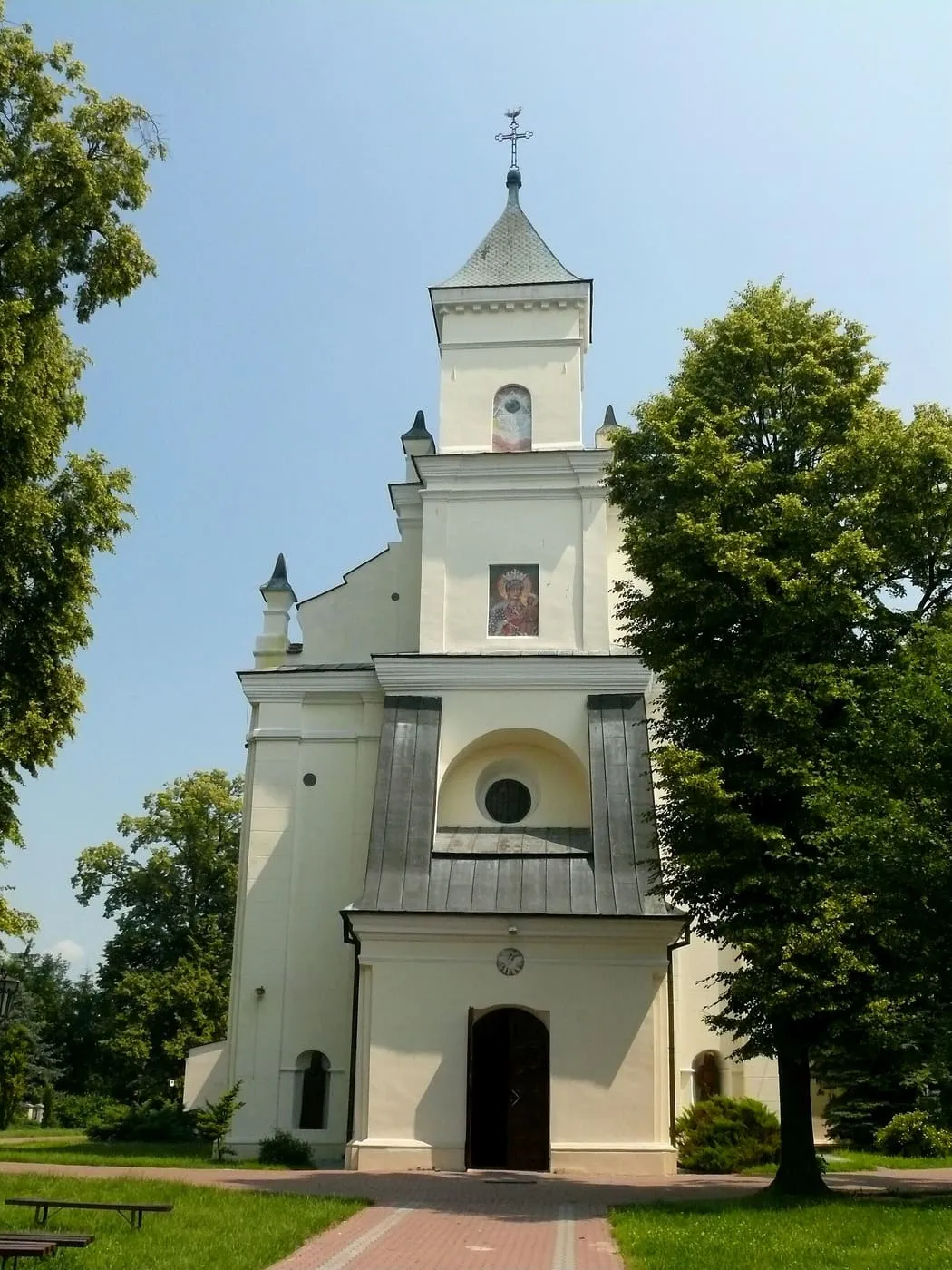 Biały kościół z wieżą na osi fasady, szarym blaszanym dachem i obrazem nad wejściem, za drzewami, z brukowaną alejką prowadzącą do drzwi
