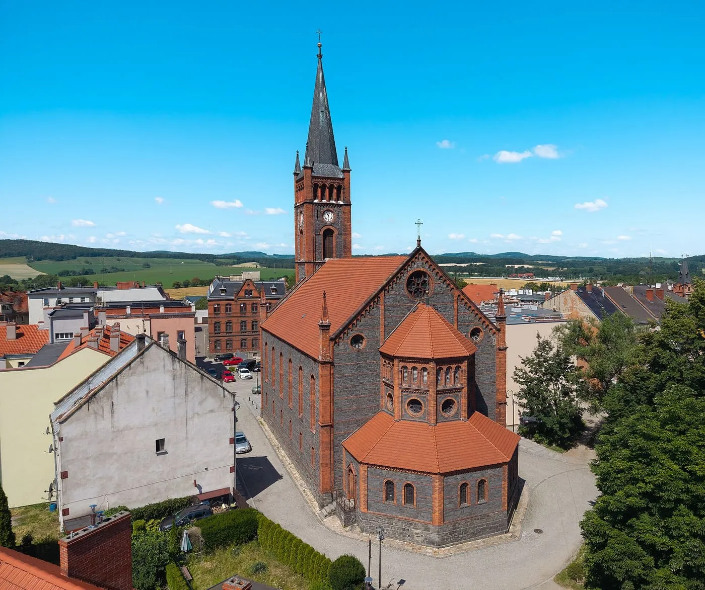 Widok z drona na ceglano-kamienny kościół z wysoką wieżą zwieńczoną iglicą i ośmiobocznym prezbiterium o czerwonych dachach, wśród zabudowy miasteczka i pagórków w tle