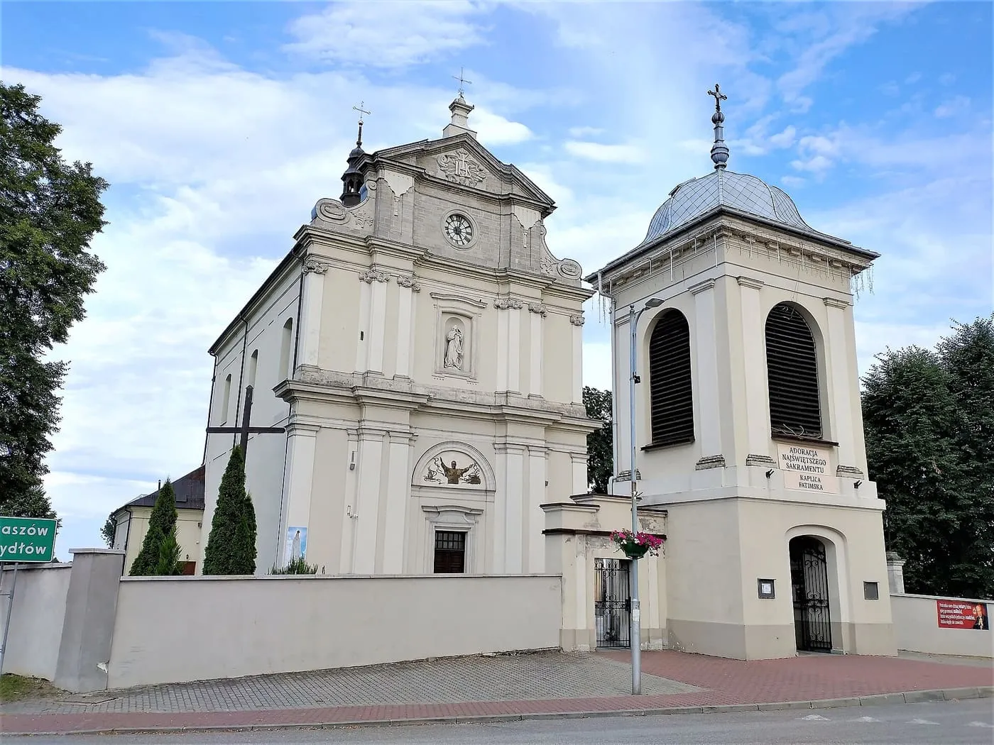 Biała, barokowa fasada kościoła z zegarem i trójkątnym szczytem oraz osobna dzwonnica z kopulastym hełmem i łukowym przejściem, za białym murem