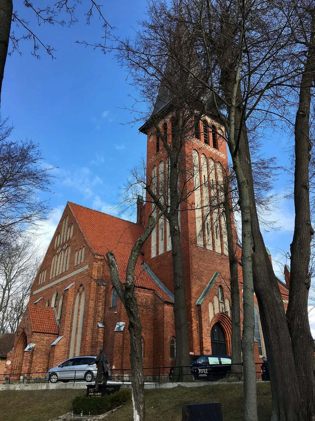 Ceglana, neogotycka wieża kościoła Niepokalanego Poczęcia w Ostródzie z kamiennymi pasami i spiczastym hełmem, widziana przez bezlistne drzewa