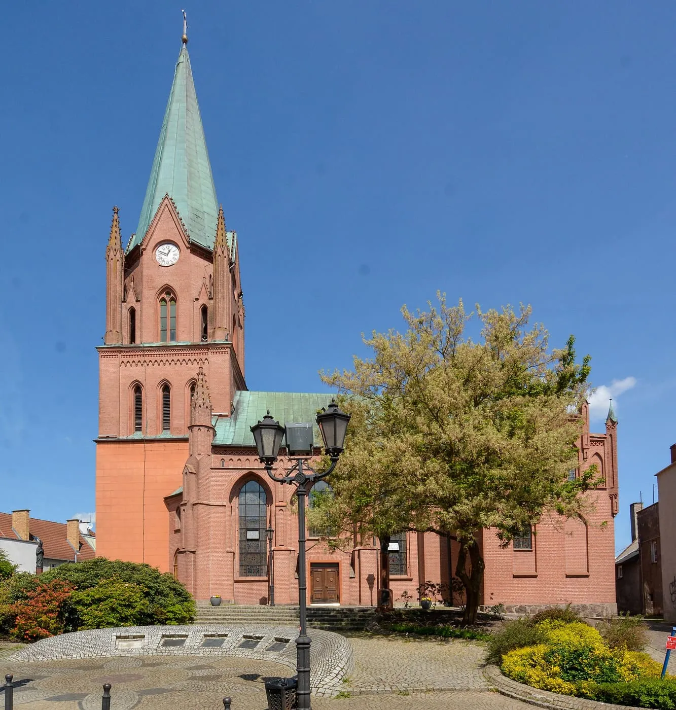 Neogotycki kościół z różowej cegły z wysoką wieżą z zegarem i zielonym hełmem, widziany z placu z latarnią, drzewem i kwietnikami