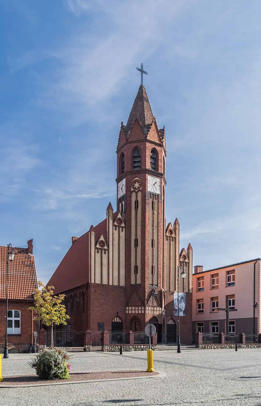 Ceglany, neogotycki kościół z wysoką wieżą z zegarem, przy wybrukowanym placu miejskim