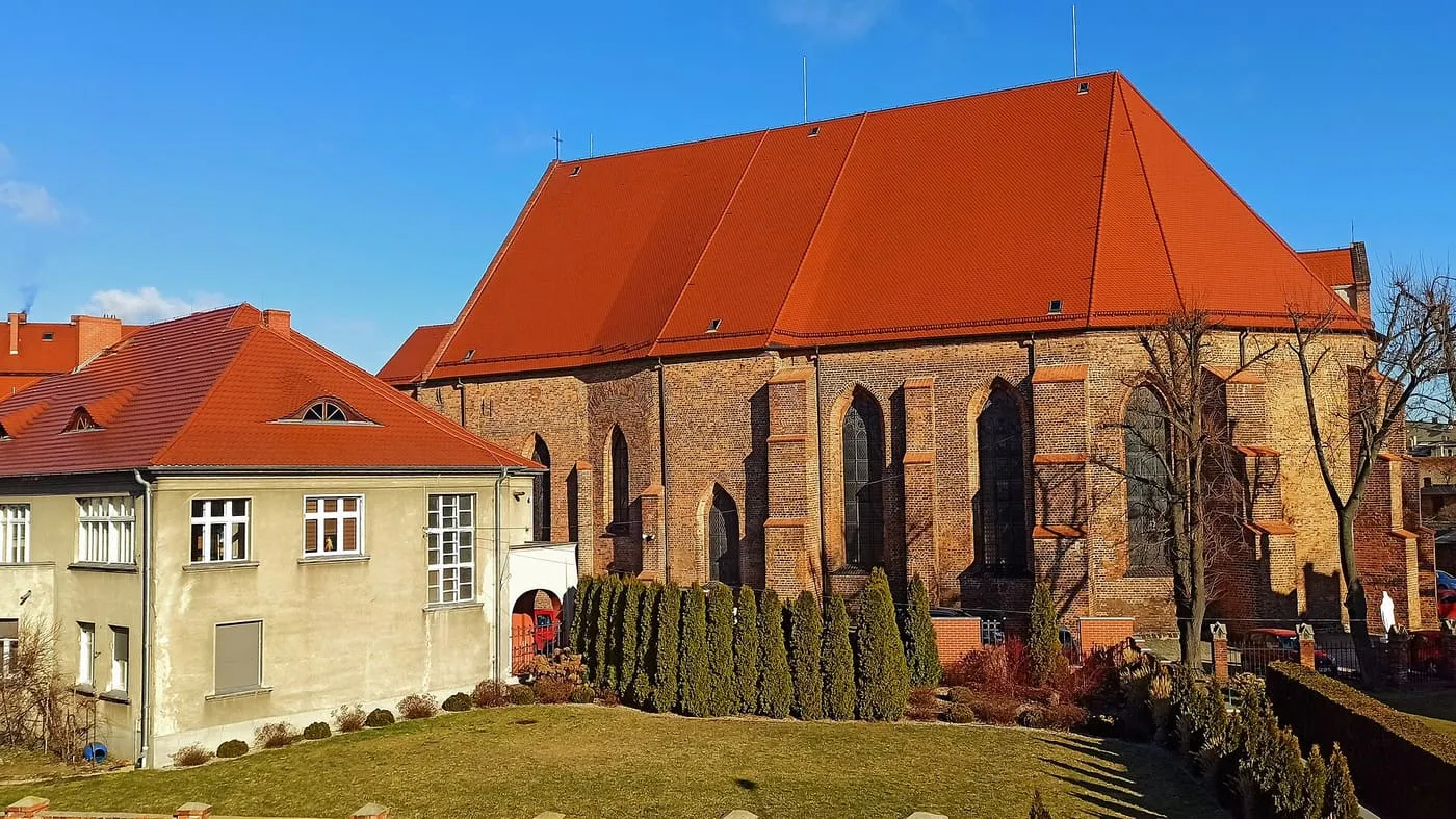 Gotycka, ceglana fara w Kościanie widziana z boku, z ogromnym czerwonym dachem, przyporami i wysokimi ostrołukowymi oknami, ponad rzędem tui, obok otynkowany budynek
