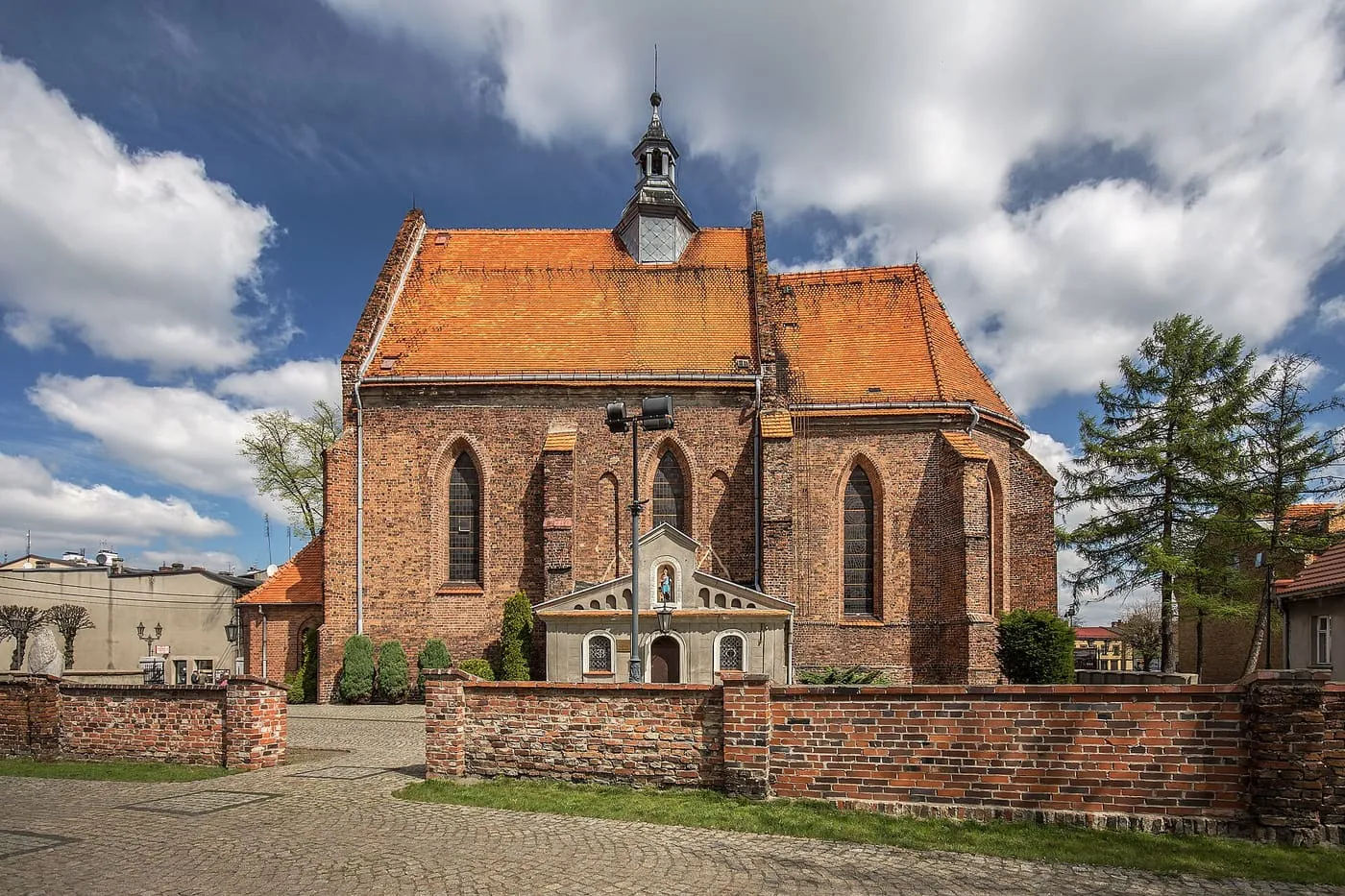 Gotycki kościół z czerwonej cegły ze stromym, pomarańczowym dachem i małą kruchtą, przed nim ceglany mur