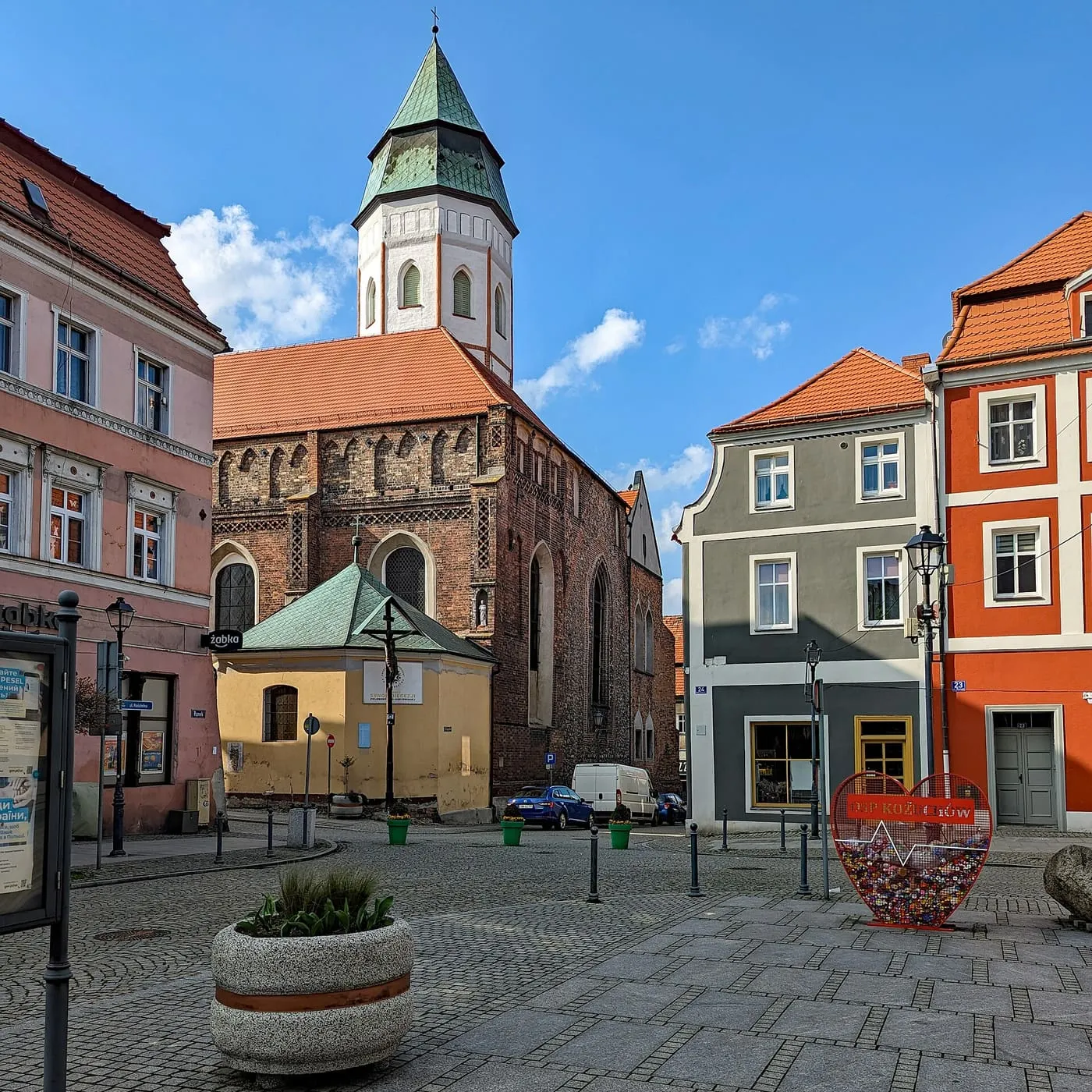 Widok z rynku w Kożuchowie na ośmioboczną wieżę fary z zielonym, namiotowym hełmem oraz ceglaną, gotycką bryłę kościoła Oczyszczenia NMP z niewielką kaplicą przy wejściu