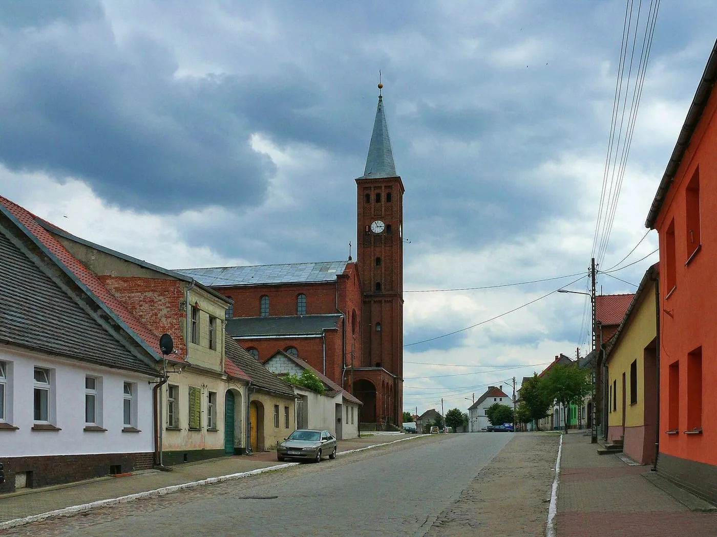 Ceglany kościół z wysoką wieżą z zegarem i smukłym hełmem, widoczny w perspektywie ulicy z kolorowymi domami