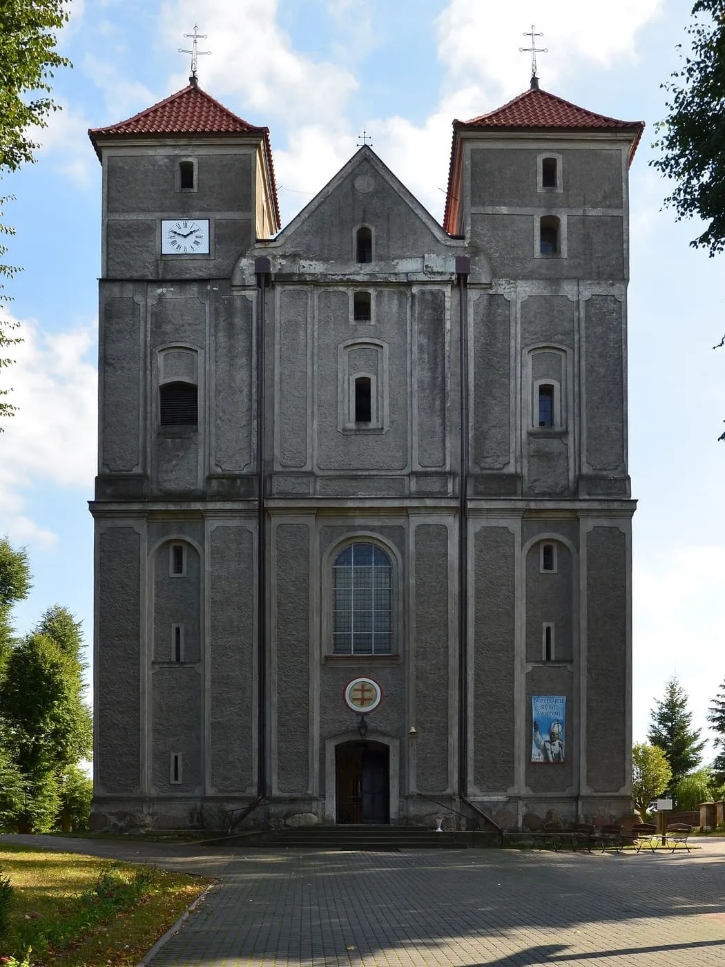 Szara, dwuwieżowa fasada kościoła z czerwonymi dachami wież, zegarem i krzyżami, przed nią wybrukowany plac