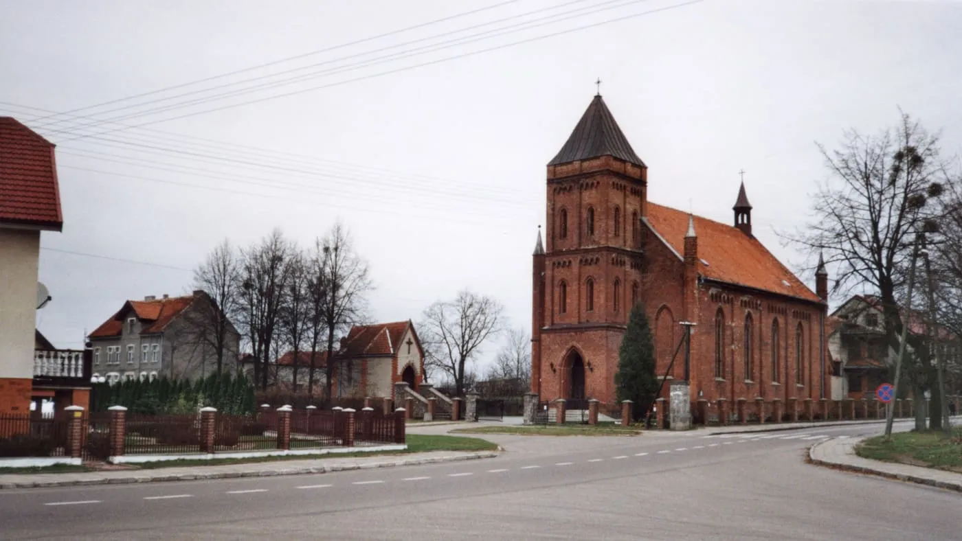 Ceglany kościół z kwadratową wieżą pod ostrym hełmem i czerwonym dachem, przy ulicy z ogrodzeniem i domami