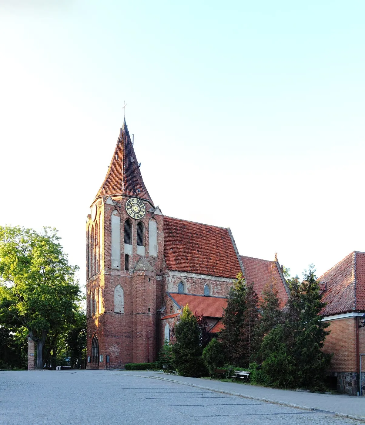 Gotycki, ceglany kościół z wieżą z zegarem i wysokim, ostrym hełmem, widziany od strony brukowanego placu, na tle jasnego nieba