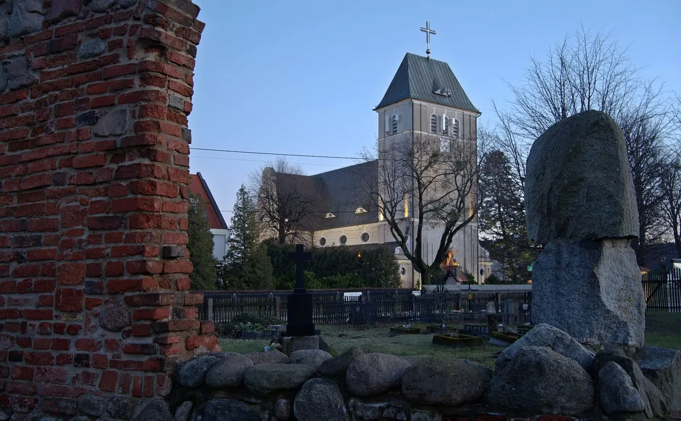 Kościół z wieżą pod ostrosłupowym dachem widoczny zza kamiennych głazów i ceglanego muru, wśród drzew bez liści