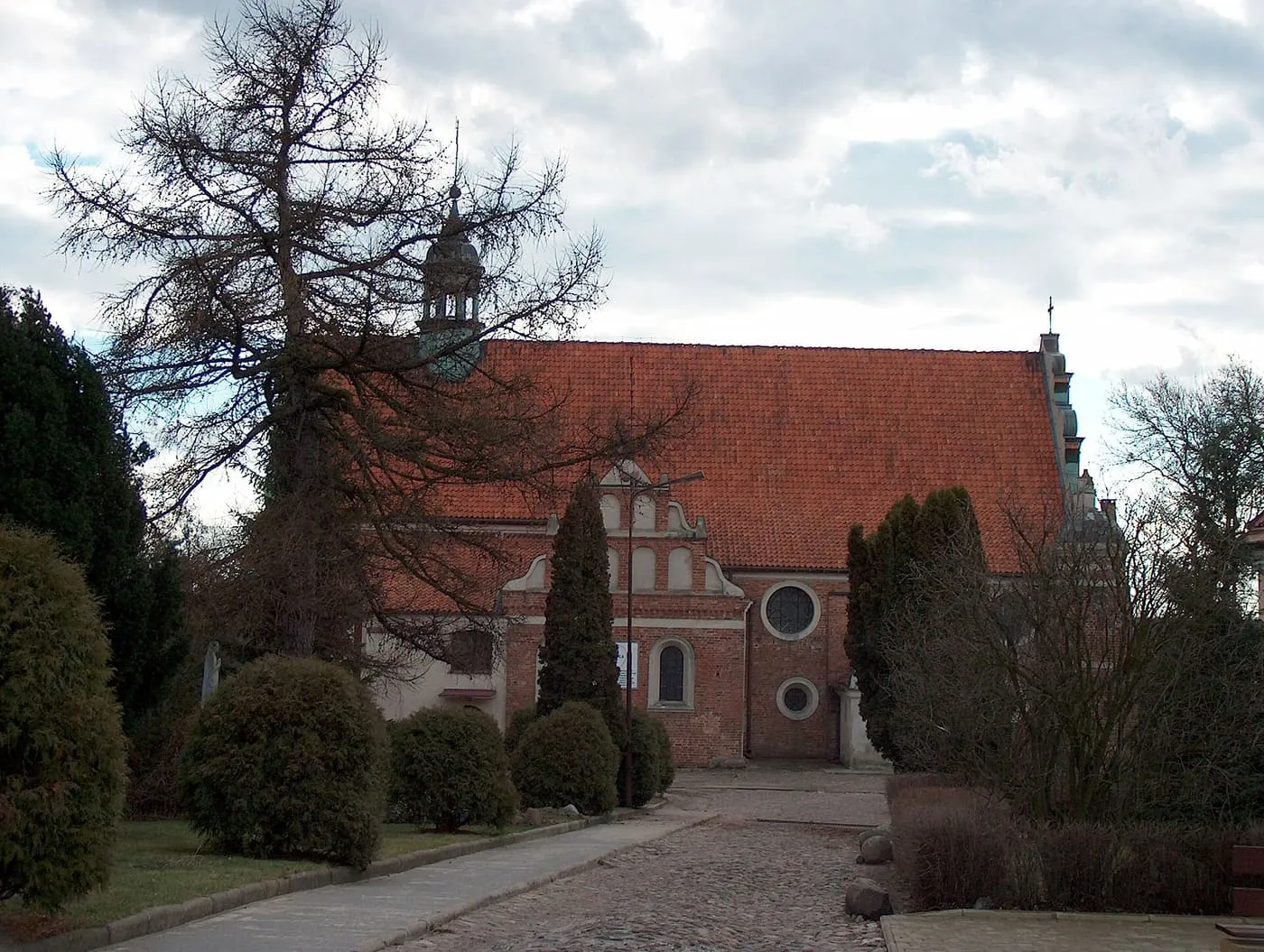 Ceglany kościół z czerwonym dachem, renesansowym szczytem i wieżyczką na sygnaturkę, częściowo zasłonięty drzewami i krzewami przy brukowanej alejce