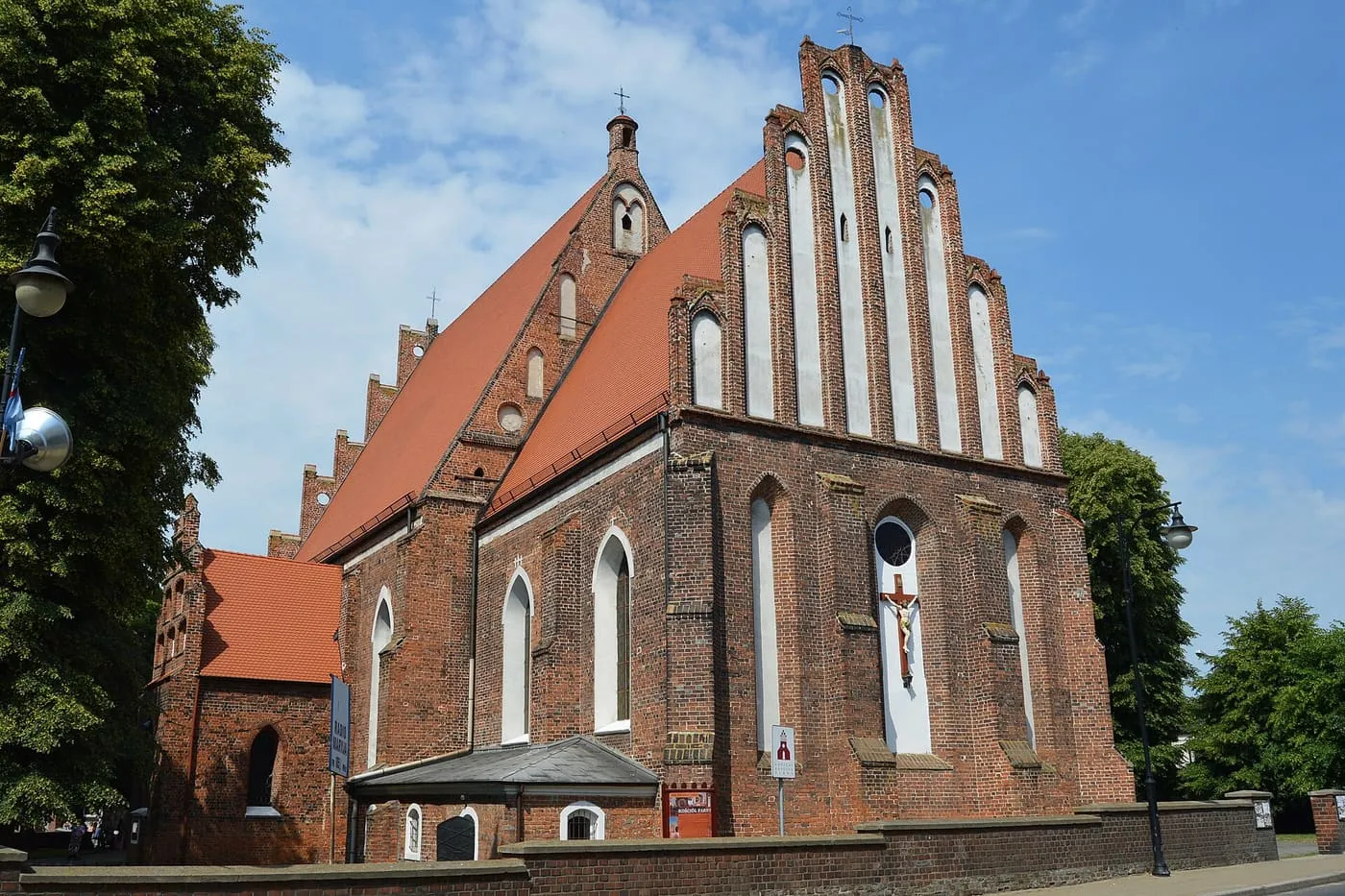 Gotycka, ceglana fara z bogato zdobionym, schodkowym szczytem z wąskimi blendami i czerwonymi dachami, z krucyfiksem w wysokiej wnęce na ścianie