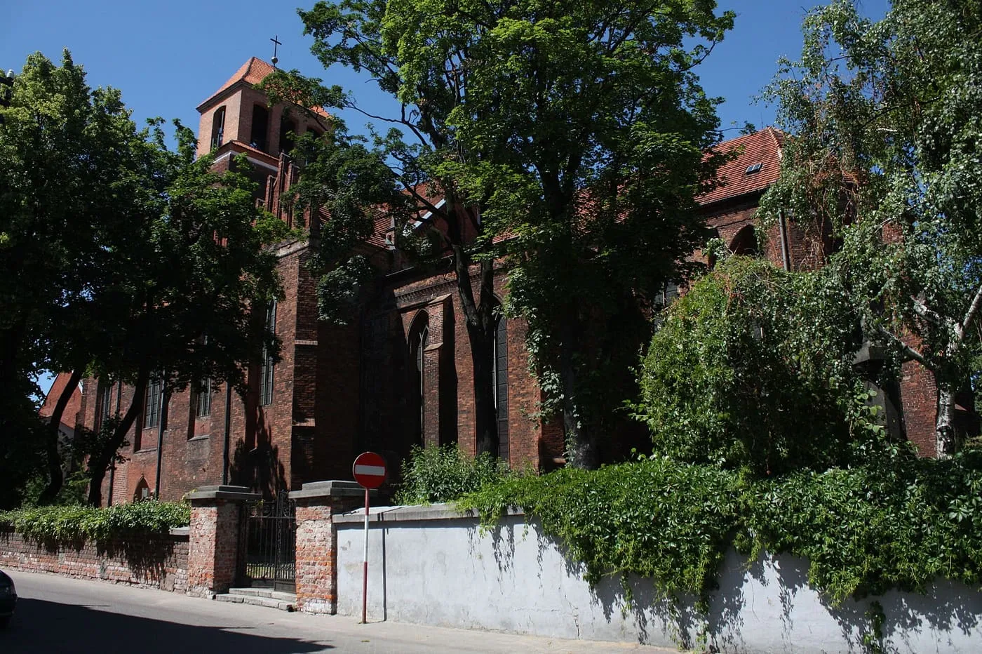 Ceglany kościół farny w Tczewie z wieżą częściowo zasłoniętą drzewami, na tle błękitnego nieba