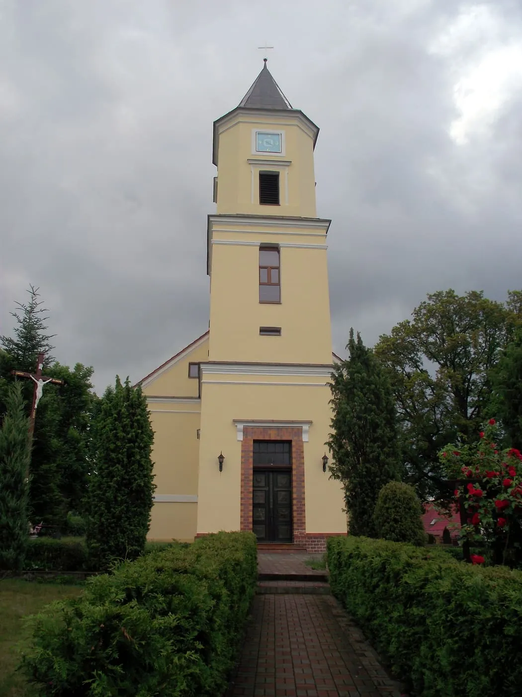 Jasnożółty, późnoklasycystyczny kościół Podwyższenia Krzyża Świętego w Torzymiu z kwadratową wieżą zwieńczoną iglicą, widziany alejką między żywopłotami