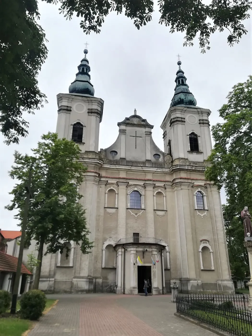 Barokowa, kremowa fasada z dwiema wieżami zwieńczonymi zielonymi cebulastymi hełmami, po prawej figura franciszkanina