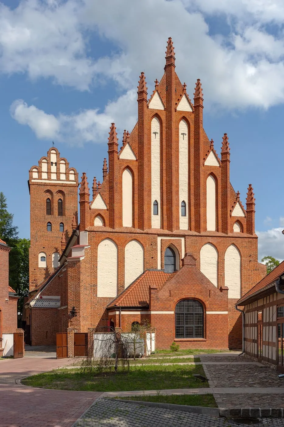 Ceglana, gotycka fasada z wysokim szczytem ozdobionym białymi blendami i fialami oraz wieżą ze schodkowym szczytem po lewej, obok trawnik i budynek szachulcowy, na tle chmur