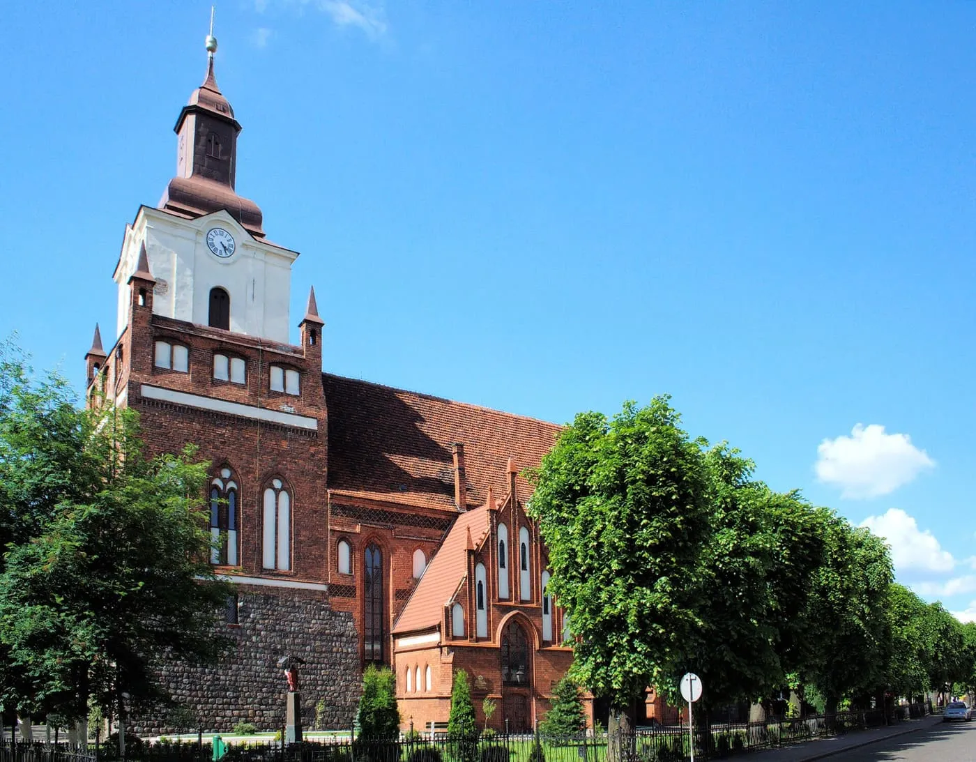Ceglany kościół z wysoką wieżą, w górnej części białą, z zegarem i ciemnym hełmem; u podstawy kamienny cokół, wokół zielone drzewa, na tle intensywnie niebieskiego nieba