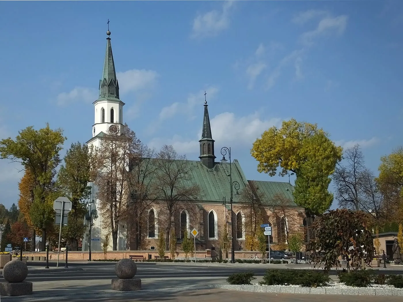 Kościół z czerwonej cegły z ostrołukowymi oknami i zielonym dachem, z białą wieżą z wysoką iglicą; wokół jesienne drzewa i skwer z kamiennymi kulami