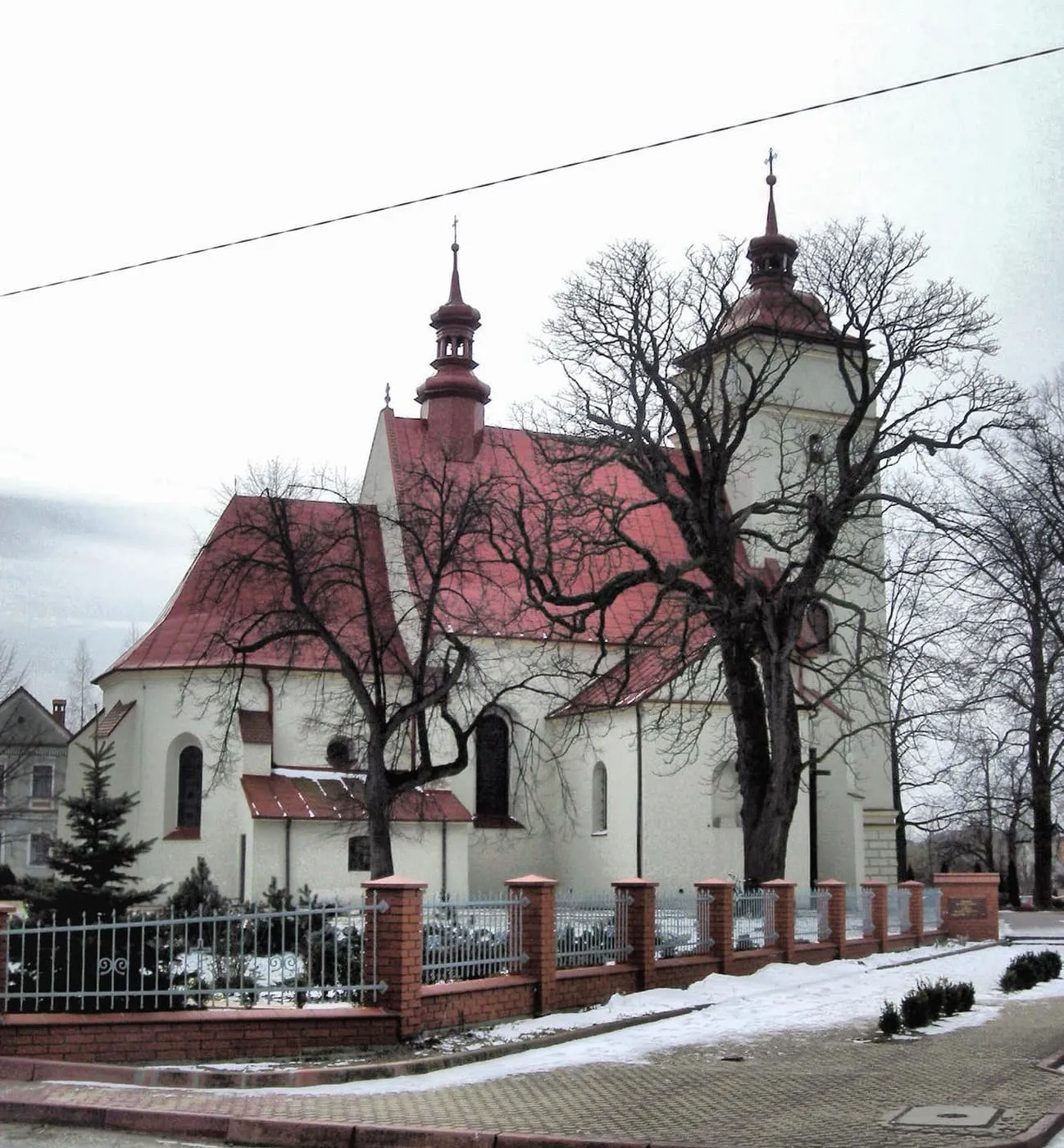 Biały kościół z czerwonymi dachami i wieżą z barokowym hełmem, otoczony bezlistnymi drzewami i ceglanym ogrodzeniem, przy lekko zaśnieżonym chodniku