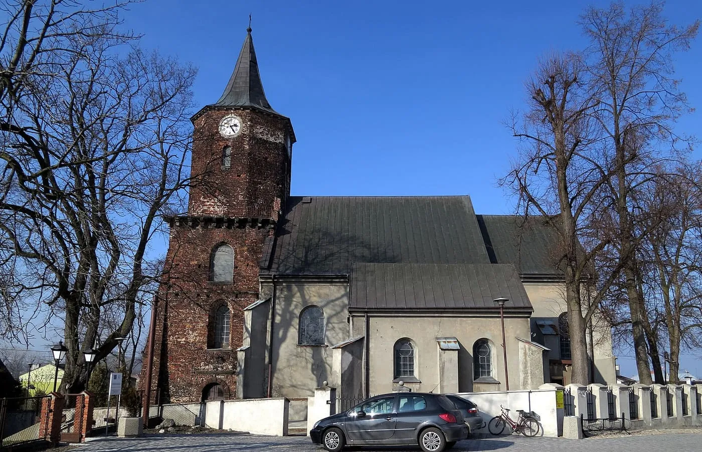 Kościół z ceglaną wieżą z zegarem i ostrym hełmem oraz ciemnym dachem nawy, przed nim biały mur i zaparkowany samochód
