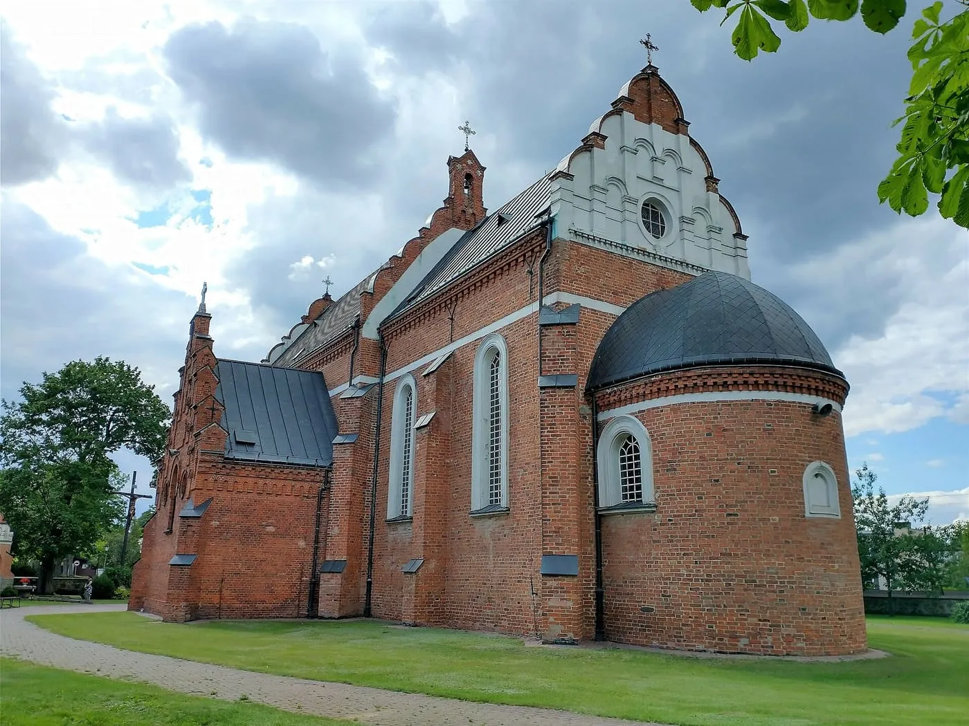 Ceglany kościół z gotyckim, schodkowym szczytem i półkolistą apsydą pod ciemną kopułą, widziany z boku na tle pochmurnego nieba