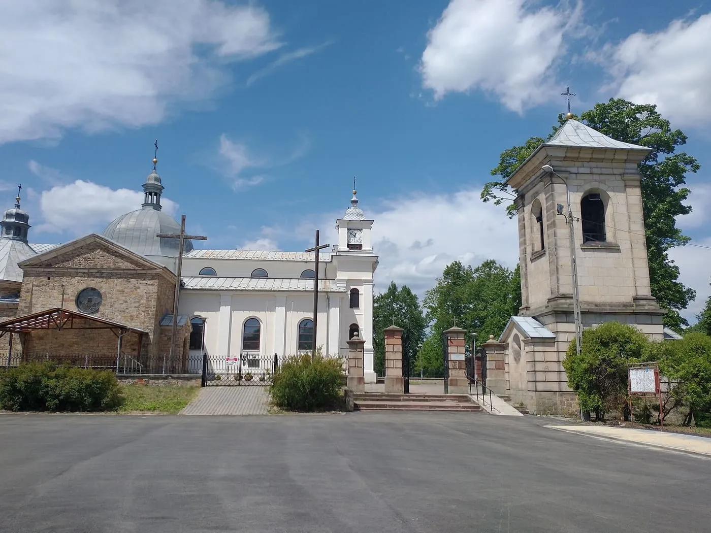 Jasny, klasycystyczny kościół z kopulastym dachem i osobną, wysoką dzwonnicą z zegarem, widziane z pustego, brukowanego placu, na tle pochmurnego nieba