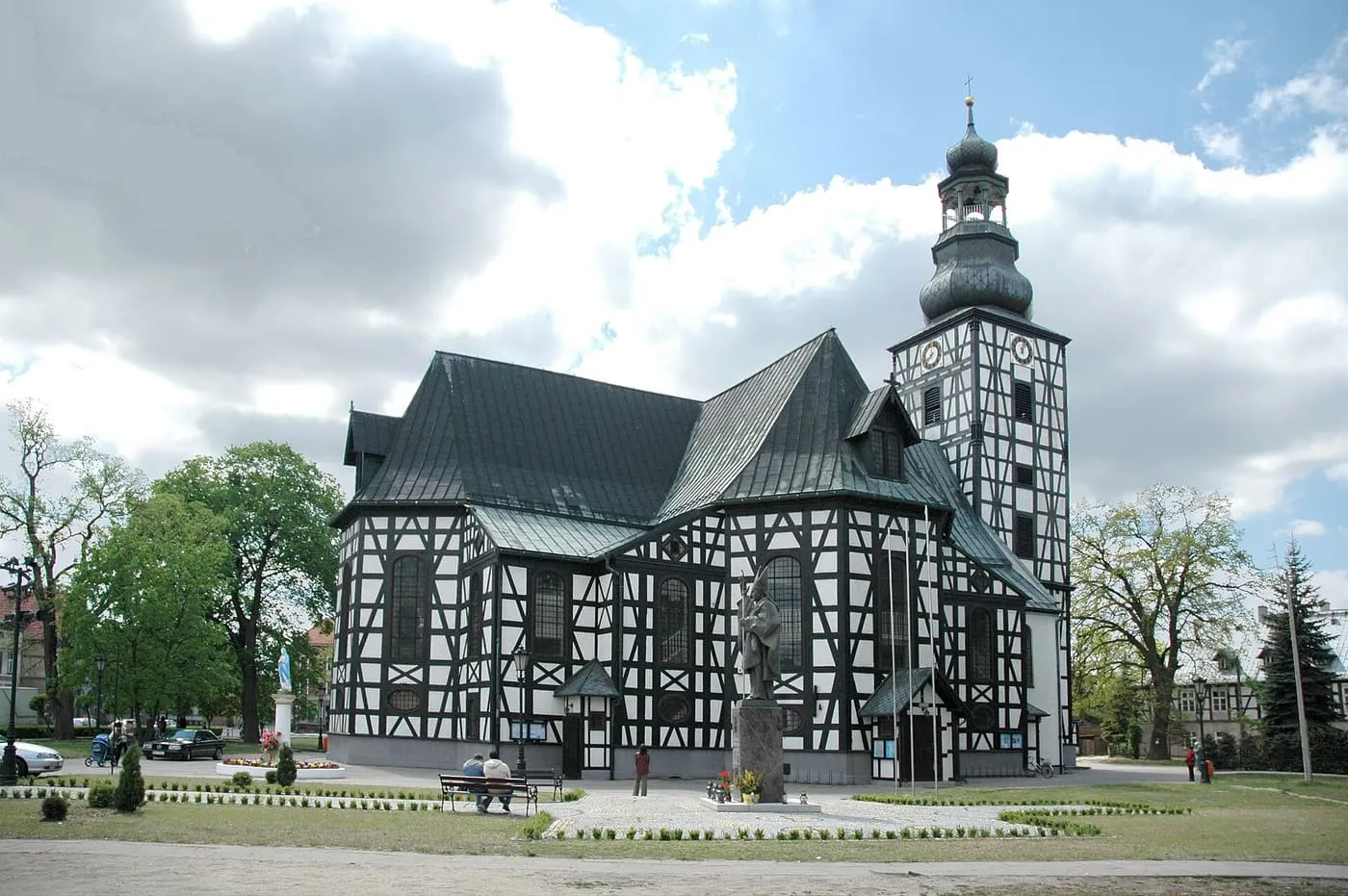 Ciemny kościół szachulcowy z jasnym wypełnieniem ścian i wysoką wieżą z cebulastym hełmem, widziany od strony zieleńca przy rynku, na tle jasnych chmur