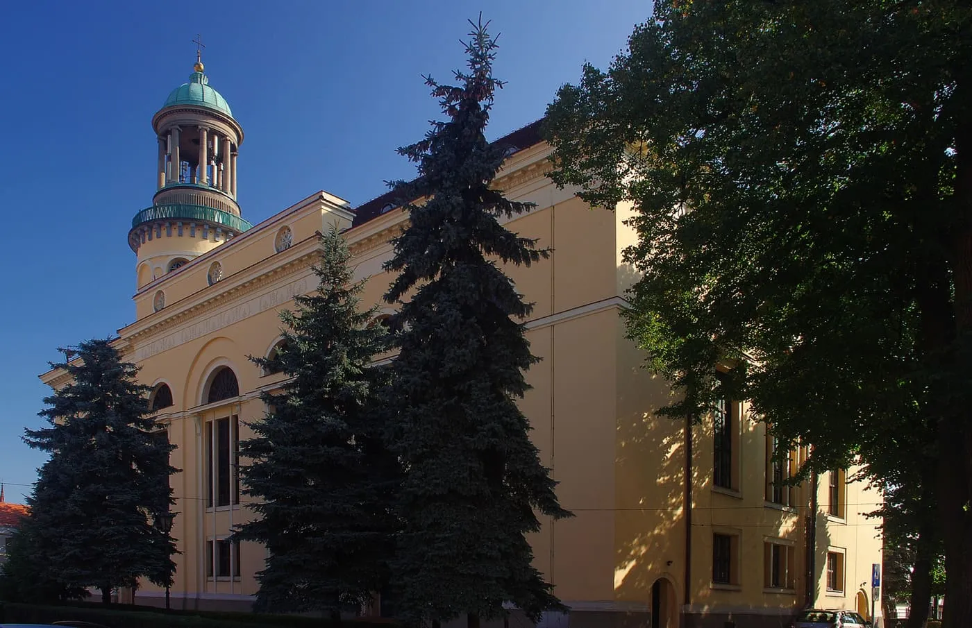 Żółty, klasycystyczny kościół z okrągłą latarnią zwieńczoną zieloną kopułą, częściowo zasłonięty ciemnymi świerkami i liściastymi drzewami, na tle granatowego nieba