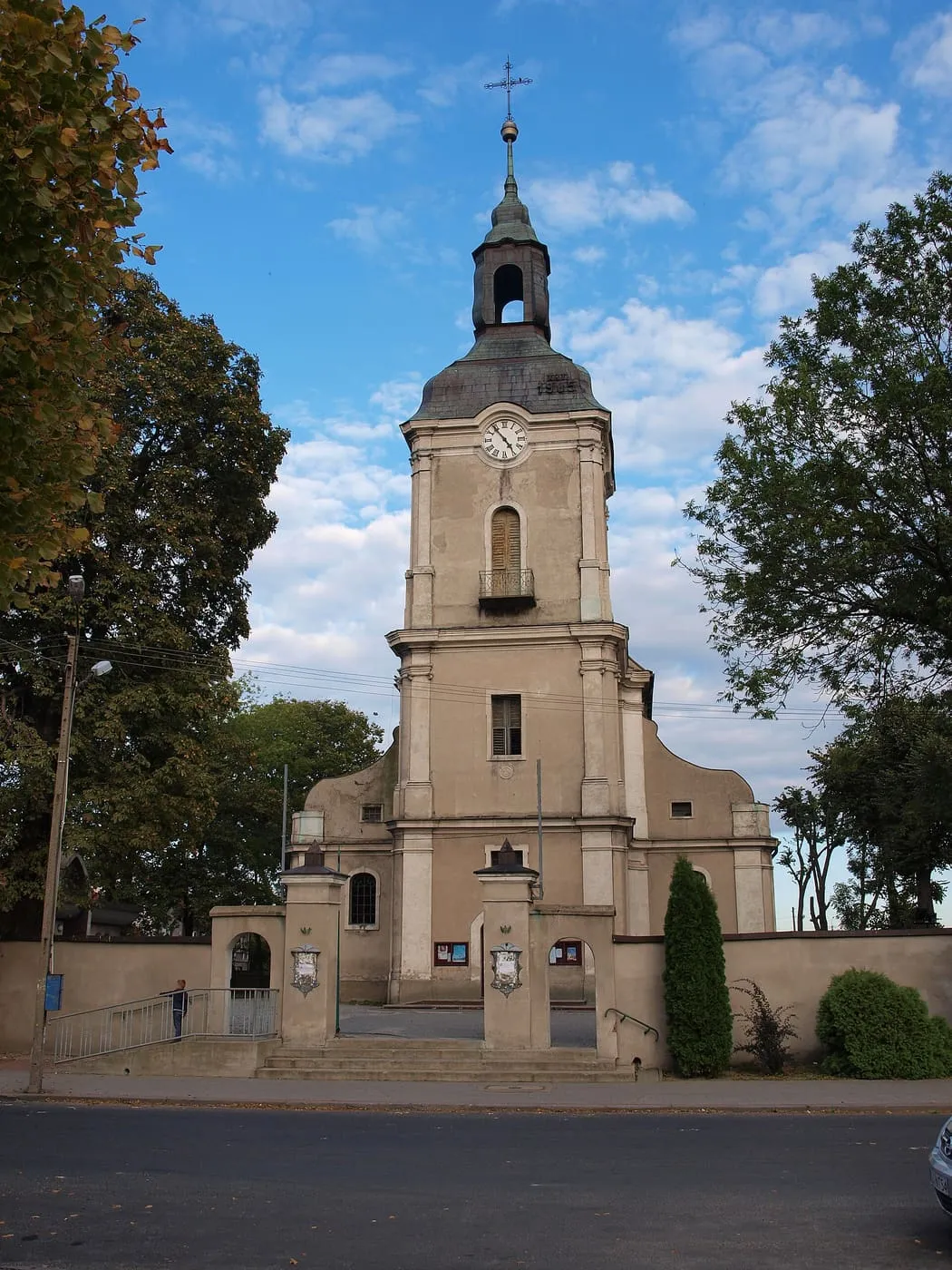 Jasna, barokowa fasada kościoła z wysoką wieżą z zegarem i ciemnym hełmem, otoczona murem, drzewami i schodami prowadzącymi do wejścia, na tle chmur