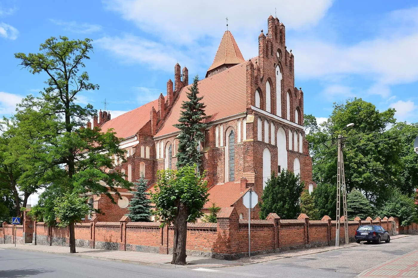 Gotycki, ceglany kościół św. Anny w Radzyniu Chełmińskim ze schodkowymi szczytami zdobionymi białymi blendami i wieżą nakrytą ostrosłupowym dachem z czerwonej dachówki, za ceglanym murem