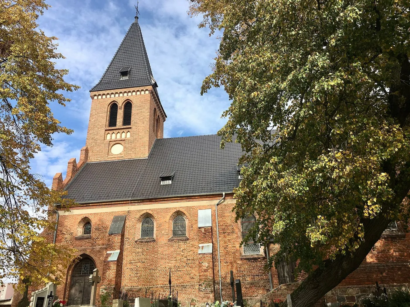 Ceglany kościół z niską, czworoboczną wieżą z ciemnym, spiczastym hełmem i dwuspadowym dachem, w jesiennej scenerii, pod koronami drzew z żółknącymi liśćmi