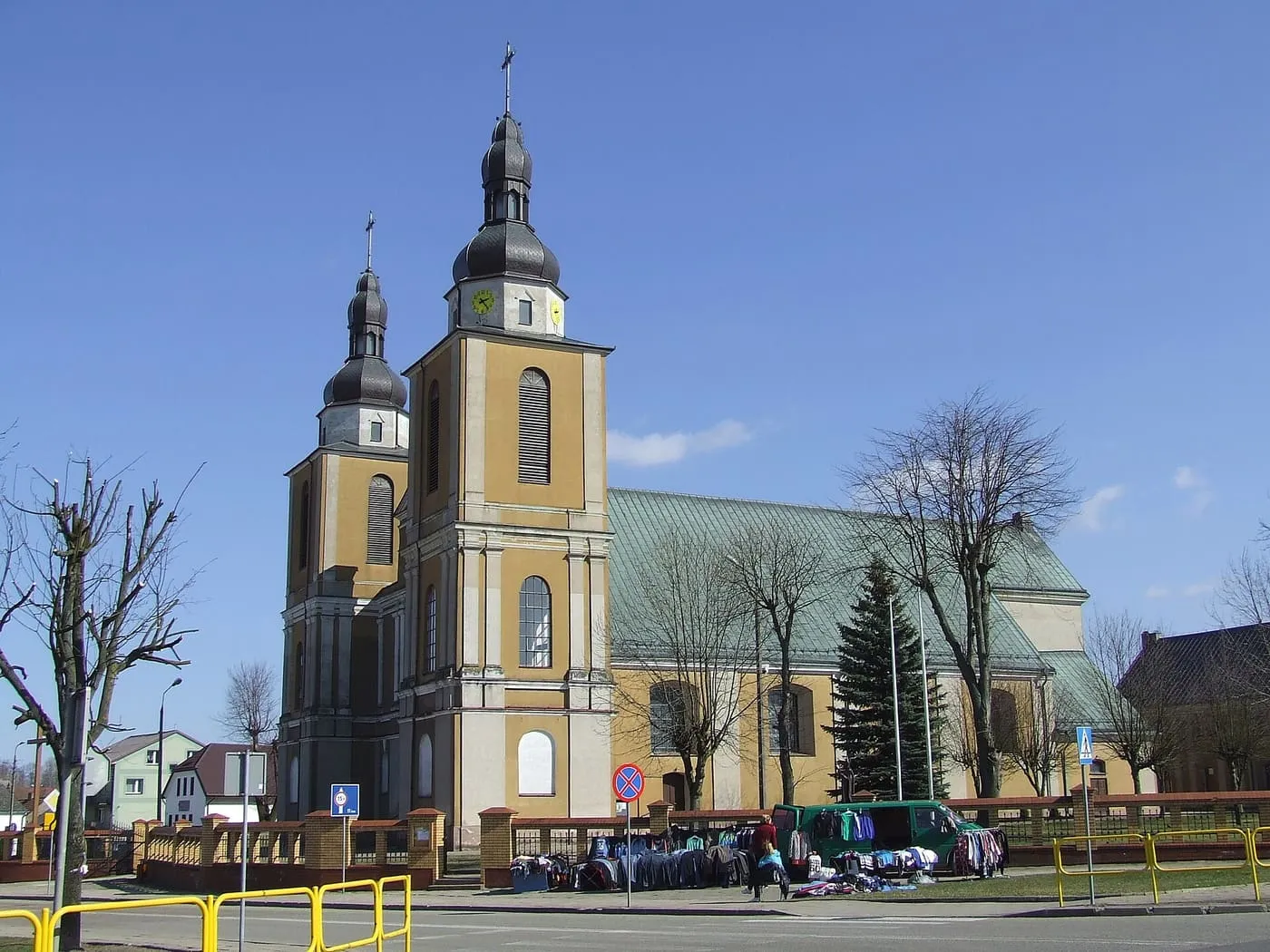 Żółta, barokowa fasada z dwiema wieżami o ciemnych hełmach i zielonym dachem nawy, widziana z ulicy, przed nią ogrodzenie i stoiska z odzieżą, na tle błękitnego nieba