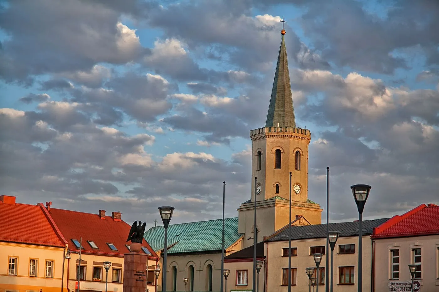 Wieża kościoła z zegarami i wysokim, zielonkawym hełmem wznosi się nad rynkiem, otoczona kolorowymi kamienicami i latarniami, na tle chmur oświetlonych zachodzącym słońcem